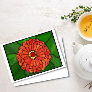 以下が含まれることがあります： 鮮やかなオレンジと黄色の花の模様が描かれたグリーンの葉のグリーティングカード。白い表面に置かれ、白いティーポットとティーカップの隣にあります。新鮮なハーブも表示されています。