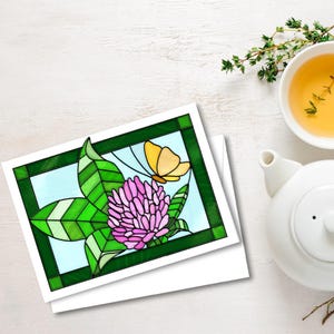 以下が含まれることがあります： ピンクの花、緑の葉、黄色の蝶が描かれたステンドグラスのデザインの白いカードが2枚。デザインは緑色で縁取られています。白いティーポットと、タイムの小枝が入ったお茶のカップが背景にあります。