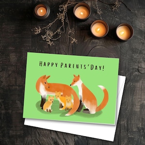 May include: A greeting card with the text "HAPPY PARENTS' DAY!" features a family of foxes on a green background. The card is on a dark wooden surface with lit candles and decorative sprigs.