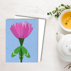 以下が含まれることがあります： 明るい青色の背景に、ピンクの花のイラスト、緑色の茎と葉が描かれたグリーティングカード。カードの後ろには白い封筒があります。白いティーポットとティーカップが右側にあります。