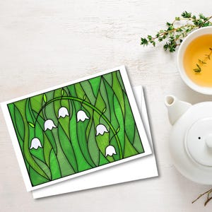 以下が含まれることがあります： 白いスズランの花と緑の葉のステンドグラス風イラストが描かれたグリーティングカード。カードは白い表面に置かれ、白いティーポットと紅茶の入ったカップの隣にあります。