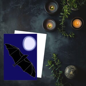 以下が含まれることがあります： 暗い青色の背景に満月を背景に飛んでいる黒いコウモリのシルエットが描かれたグリーティングカード。カードは白い封筒と組み合わされています。シーンは、ろうそくと緑のある暗い表面に設定されています。