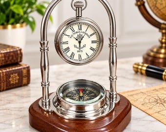 May include: A silver-toned metal desk clock and compass set mounted on a dark wood base. The clock face features Roman numerals, the text "VICTORIA LONDON 1875," and a ship's wheel and anchor graphic. The compass sits below the clock.