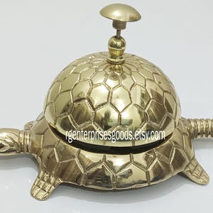 May include: A gold-toned brass turtle-shaped service bell with a hexagonal patterned top. The bell has a small knob on top and a coiled tail.