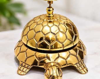 May include: A polished gold-tone metal service bell shaped like a turtle. The dome of the bell features a textured hexagonal pattern resembling a shell, with a small circular knob at the top. The base includes a turtle head and four feet.
