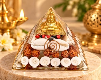 May include: A clear resin pyramid containing a gold-colored temple structure, white cowrie shells, brown textured rudraksha beads, small red and black seeds, and a copper wire spiral. The pyramid is placed on a circular stone base.