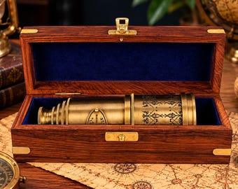 May include: A brass nautical telescope with intricate black etched patterns rests inside a rectangular wooden storage box. The box features a dark wood grain finish, brass corner accents, a central brass latch, and a deep blue velvet interior lining.
