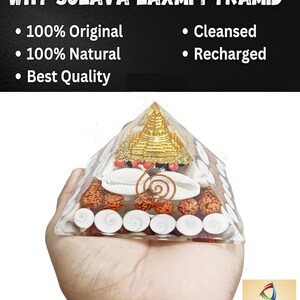 May include: A white crystal pyramid with gold accents and red beads. The pyramid is filled with white seashells, brown beads, and a copper wire spiral. The pyramid is being held in a person's hand. The text "WHY SOLAVA LAXMI PYRAMID" is at the top of the image. The text "100% Original", "100% Natural", "Best Quality", "Cleansed", and "Recharged" are listed below the pyramid. The text "RG MART" is in the bottom right corner of the image.