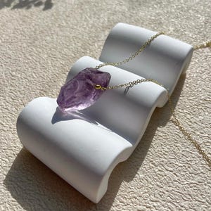 May include: A delicate gold chain necklace with a large, raw amethyst pendant. The amethyst is a deep purple colour and has a natural, irregular shape.