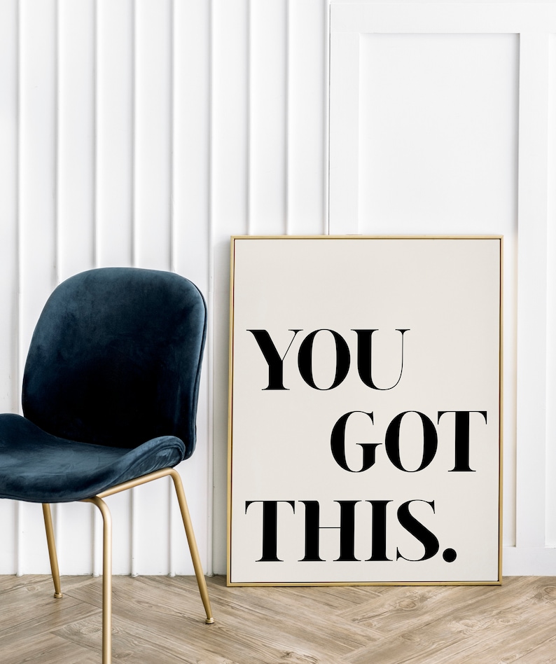 May include: A motivational poster with a gold frame, featuring the text "YOU GOT THIS." in black bold font on a cream background. The poster is leaning against a white wall next to a blue velvet chair with gold legs.