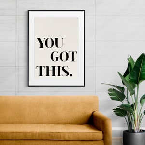 May include: A black framed print with the text "YOU GOT THIS." on a white background. The print is hanging on a white tiled wall above a brown couch.