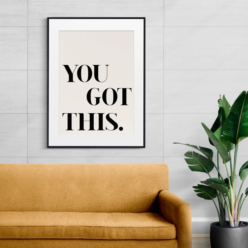 May include: A black framed print with the text "YOU GOT THIS." on a white background. The print is hanging on a white tiled wall above a brown couch.