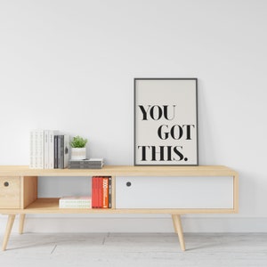 May include: A black framed poster with the text "YOU GOT THIS." on a white background. The poster is hanging on a white wall above a light wood entertainment center with books and a potted plant.