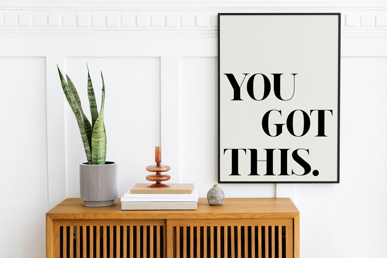 May include: A framed motivational poster with the text "YOU GOT THIS." on a white background. The poster is hanging on a white wall above a wooden cabinet with a potted snake plant, a stack of books, and a decorative candle holder.