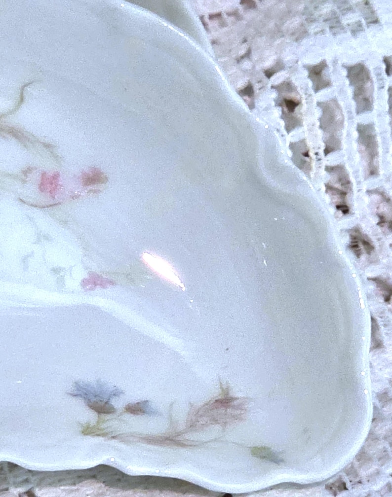 May include: A white porcelain dish with a scalloped edge. Delicate floral designs in pink, blue, and brown adorn the surface. The dish has a glossy finish and appears to be vintage or antique.
