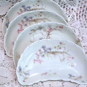 May include: Four white, scalloped-edge porcelain plates. Each plate features a delicate floral design in pink, blue, and green. The floral pattern is arranged in a graceful, flowing style across the plates' surfaces. The plates are arranged in a slightly overlapping manner.