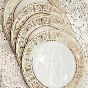 May include: A stack of white porcelain plates with gold filigree detailing. The plates have a wide gold border with an ornate, repeating pattern. The center of each plate is plain white. The plates are arranged on a white lace tablecloth.