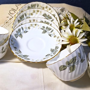 May include: A set of white porcelain teacups and saucers featuring a delicate green leaf and small flower pattern with gold-trimmed scalloped edges. The cups have a ribbed texture and curved handles, arranged with a floral accent piece.