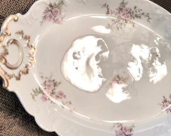 Op de afbeelding: Ovale, witte porseleinen serveerschaal met een geschulpte rand en gouden accenten. De schaal is versierd met roze bloemmotieven. De handvatten hebben een gouden ontwerp. Een decoratief stuk voor het serveren van eten.