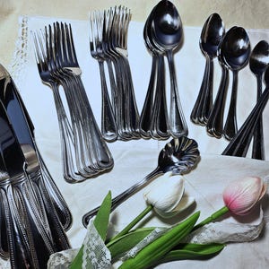 May include: A collection of polished silverware, including forks, spoons, and knives, arranged on a white cloth. The silverware has a decorative beaded edge. Two tulips, one white and one pink, are placed near the silverware.