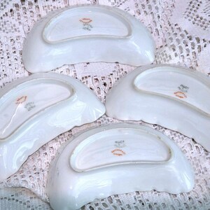 May include: Four white porcelain dishes with a scalloped edge. Each dish has a curved, crescent shape and a decorative emblem in the centre. The dishes are arranged on a white lace surface.