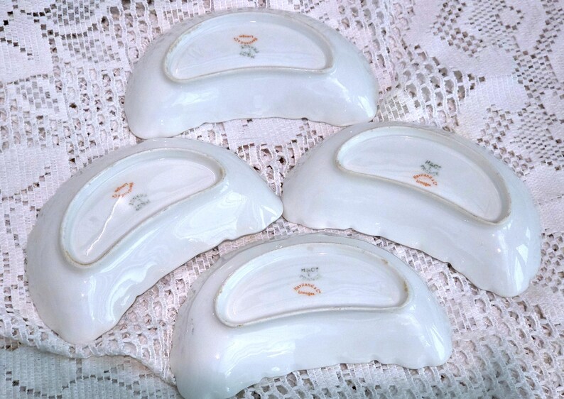 May include: Four white porcelain dishes with a scalloped edge. Each dish has a curved, crescent shape and a decorative emblem in the centre. The dishes are arranged on a white lace surface.