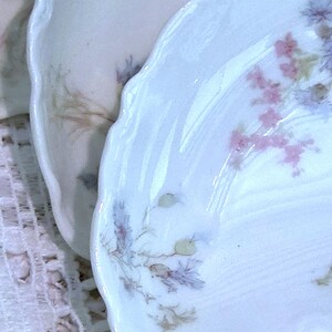 May include: Close-up of antique porcelain plates with scalloped edges. The plates feature delicate floral patterns in pink, purple, and green on a white background. The plates are likely vintage and add a touch of elegance.