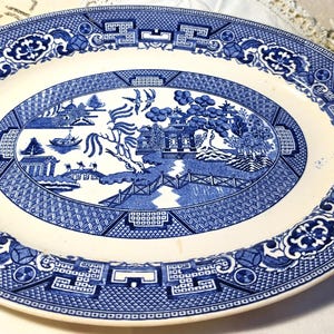 May include: An oval ceramic platter featuring a blue and white willow pattern. The central design depicts a landscape with a pagoda, a bridge, and birds, surrounded by a wide border of geometric and floral motifs on a cream-colored background.