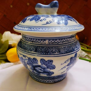May include: A white ceramic lidded jar featuring blue floral patterns and intricate geometric borders. The lid is topped with a blue-tinted handle. The jar has a rounded body and a flared base, decorated with detailed blue leaf and flower motifs.