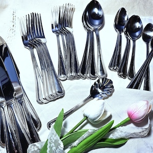 May include: A collection of stainless steel flatware including dinner knives, forks, and spoons with a beaded edge pattern on the handles. A single shell-shaped spoon is positioned near two artificial tulips with white and pink petals.