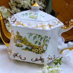 May include: A white porcelain teapot with a hexagonal shape, featuring a gold-painted spout, handle, and lid finial. The body is decorated with a colorful landscape illustration of a pagoda, willow trees, and yellow flowers, accented by gold scrollwork.