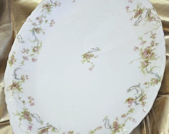 Op de afbeelding: Ovale witte serveerschaal met een bloemmotief. De schaal heeft roze en groene bloemen met blauwe lintaccenten. De rand heeft een gouden rand. Een decoratief stuk voor het serveren van eten.