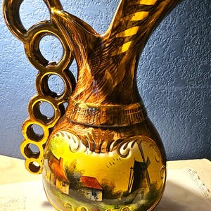 May include: A ceramic pitcher featuring a glossy brown finish with gold accents and a unique handle composed of five connected rings. The body displays a painted scene of a rural landscape with houses and a windmill, framed by decorative gold scrollwork.