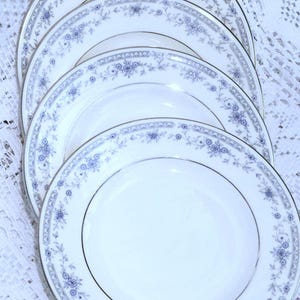 May include: A stack of white porcelain plates with a delicate blue floral pattern around the rim and a silver trim. The plates are arranged in a fan shape, showcasing the design. The plates are on a white lace tablecloth.