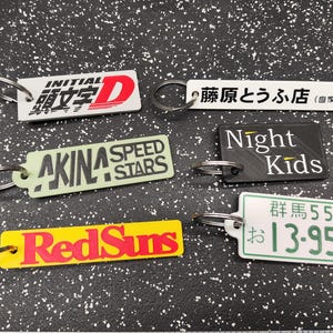 May include: Six different keychains with various designs. The keychains feature the text "INITIAL 頭文字D", "AKINA SPEED STARS", "RedSuns", "藤原とうふ店(自家用)", "Night Kids", and "群馬55 お13-954".