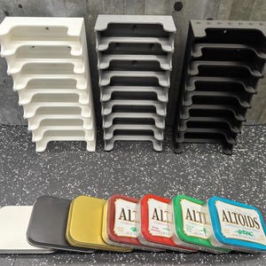 May include: Three stacks of tiered, plastic organizers in white, gray, and black. Below, a row of Altoids tins in various colors: white, black, gold, red, green, and blue. The Altoids tins have white labels.
