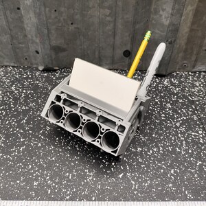 May include: A gray plastic model of an engine block with four cylinders. The model has a slot for holding a pen or pencil and a small card. A yellow pencil and a white pen are in the slots.