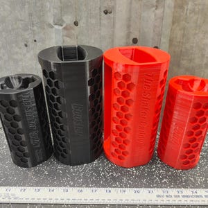 May include: Four cylindrical shaker caddies with a hexagonal honeycomb pattern on the exterior. Two are black and two are red. The black units are labeled The Shaker Caddy and Booster. The red units are labeled The Shaker Caddy and Caddy. A ruler is visible.