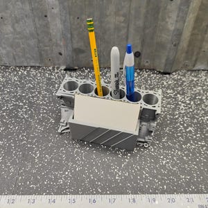 May include: A gray desk organizer shaped like an engine block with six cylindrical holes for pens and pencils, featuring a front slot holding a blank rectangular card. A yellow pencil, a gray marker, and a blue pen are inserted into the holes.