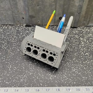 May include: A gray engine block desk organizer featuring a rectangular slot for business cards and three cylindrical openings for pens. The organizer holds a yellow pencil, a blue pen, and a white marker, positioned above a ruler showing inches.