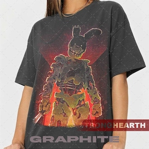 May include: Dark gray t-shirt featuring a graphic of a menacing, decaying rabbit-like figure with a knife, set against a red and orange background. The text "STRONG HEARTH" and "GRAPHITE" are printed at the bottom.