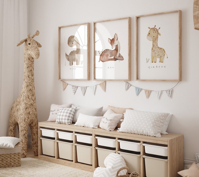 May include: A light wood shelf unit with white storage bins and three framed watercolour prints of a dinosaur, a deer, and a giraffe. The prints have the names of the animals written below them. A stuffed giraffe toy stands next to the shelf unit.
