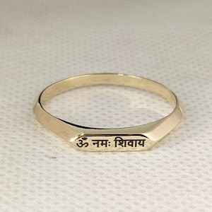 May include: A gold band ring with a flat, rectangular top engraved with the Sanskrit phrase "Om Nam: Shivaya".