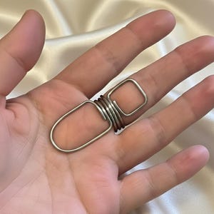 Peut inclure: Une bague argentée au design unique. La bague présente une forme rectangulaire reliée à une bande enroulée et une forme ovale. La bague est portée au doigt.