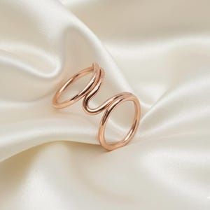 Puede incluir: Un anillo doble para dos dedos en oro rosa con un dise&ntilde;o &uacute;nico y fluido. El anillo presenta dos bandas conectadas, una para cada dedo, creando un aspecto moderno y elegante. El anillo se muestra sobre una superficie de sat&eacute;n blanco.