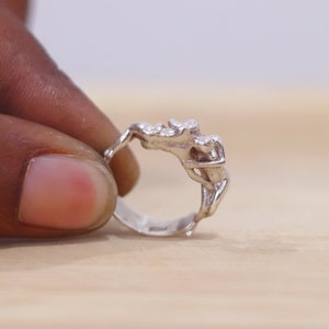 Può includere: Un anello d'argento con un designo scolpito di due figure che si abbracciano. L'anello è mostrato su un dito.