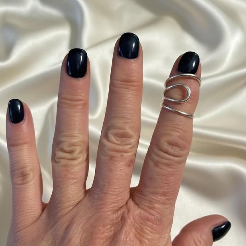 Puede incluir: Un anillo de dedo plateado con un dise&ntilde;o curvo &uacute;nico. El anillo envuelve el dedo con una est&eacute;tica minimalista y moderna. Las u&ntilde;as est&aacute;n pintadas de un color oscuro y brillante.