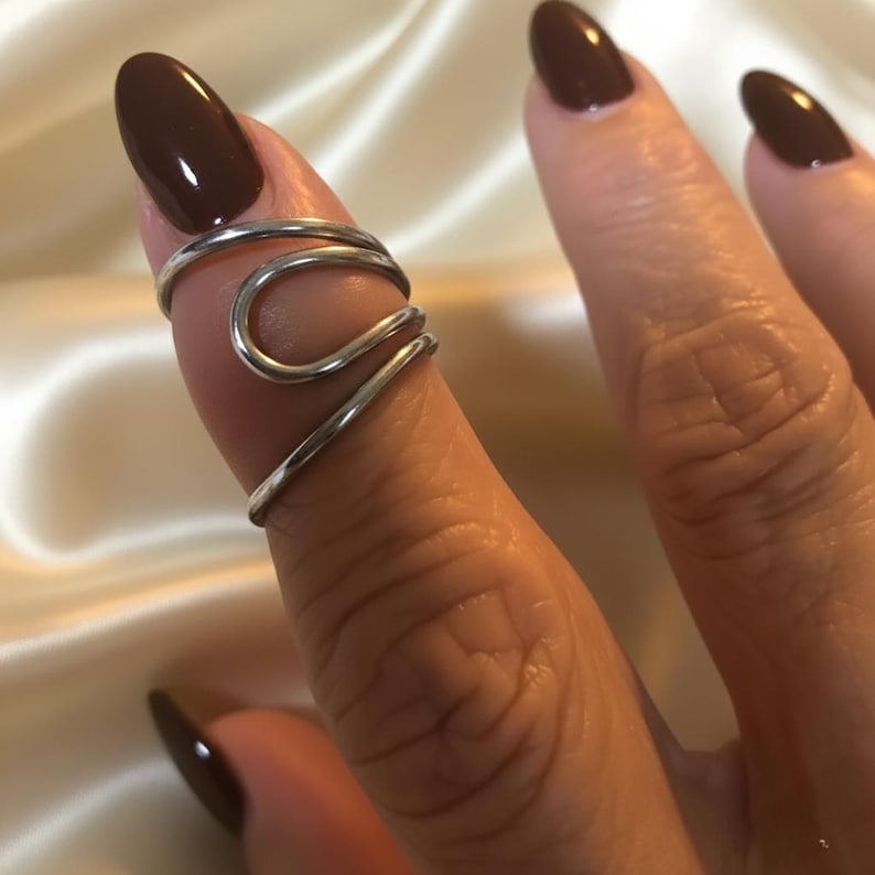 Puede incluir: Un anillo de dedo plateado con un dise&ntilde;o &uacute;nico y fluido. El anillo se envuelve alrededor del dedo con una serie de l&iacute;neas curvas, creando un aspecto abstracto y moderno. El anillo se muestra en un dedo con esmalte de u&ntilde;as marr&oacute;n oscuro.