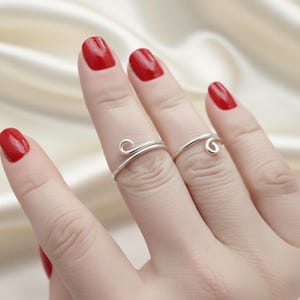 Op de afbeelding: Twee zilveren ringen met een minimalistisch ontwerp worden getoond op vingers met rode nagellak. De ringen hebben een delicaat, wervelend detail, wat elegantie toevoegt. De ringen staan tegen een zachte, neutrale achtergrond.