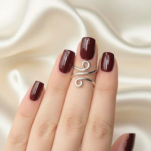 May include: A silver-colored, adjustable ring with a swirling design is worn on a finger with dark red nail polish. The ring wraps around the finger with a delicate, openwork pattern.